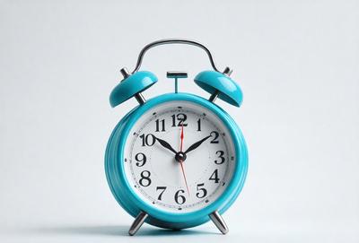 The primary subject of the image is a blue alarm clock with a white face. The clock is placed on a white surface, which serves as the setting or background. The visual style of the image is a photo, capturing the alarm clock in its entirety. The dominant colors in the image are blue and white, creating a calm and soothing atmosphere.