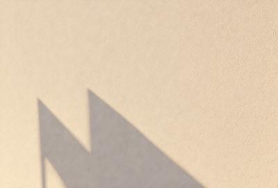 The primary subject of the image is a shadow of a triangle on a wall. The setting is a plain wall, and the visual style appears to be a photo. The colors in the image are predominantly neutral, with the shadow being the main focus. The overall atmosphere of the image is minimalistic and simple.