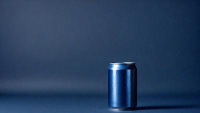 The primary subject of the image is a blue can of soda. The setting is a dark blue background, which creates a contrasting and dramatic effect. The visual style is a photo, capturing the details of the can and its surroundings. The colors and mood of the image are predominantly dark and moody, with the blue can standing out against the dark blue background.