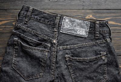 The primary subject of the image is a pair of jeans. The jeans are displayed on a wooden table, showcasing their design and texture. The setting is a casual and comfortable environment, likely a home or a store. The visual style is a close-up of the jeans, highlighting their details and quality. The colors and mood of the image are predominantly dark, with the jeans being the main focus.