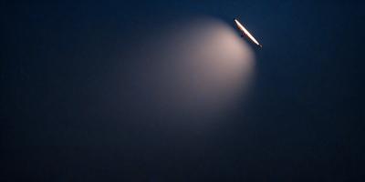 The primary subject of the image is a light source, possibly a light bulb or a lightning bolt, shining brightly in the dark. The setting is a foggy, cloudy sky, creating a moody atmosphere. The visual style is a combination of a photo and a digital art, as the light source appears to be a digital creation. The colors are predominantly dark, with the light source being the only bright element in the scene.
