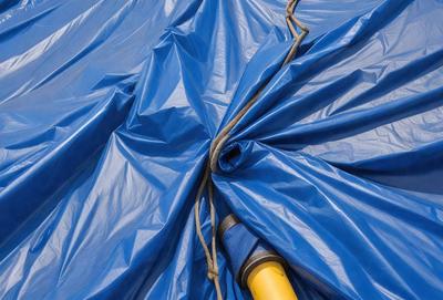 The primary subject of the image is a blue tarp with a yellow pipe sticking out of it. The setting is an outdoor environment, with the tarp covering a surface. The visual style is a photo, capturing the details of the tarp and the pipe. The colors and mood of the image are predominantly blue and yellow, creating a contrasting and vibrant atmosphere.