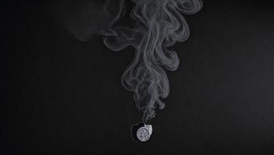 The primary subject of the image is a small, round object, possibly a clock or a bullet, floating in the air. The setting is a black background, which creates a dramatic and mysterious atmosphere. The visual style appears to be a combination of a photo and a drawing, with the object being in focus and the background being blurred. The colors in the image are predominantly black, which further emphasizes the dramatic and moody mood of the scene.