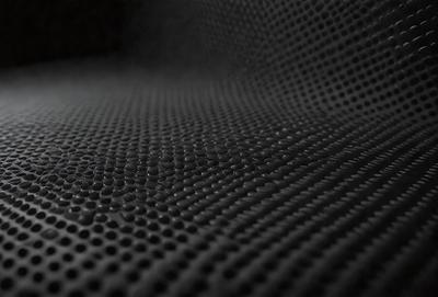 The primary subject of the image is a close-up of a black mesh material, possibly a net or a patterned surface. The setting appears to be a dark room, with the mesh material being the main focus of the image. The visual style is a close-up, which emphasizes the details of the mesh material. The colors in the image are predominantly black, giving the scene a moody and mysterious atmosphere.
