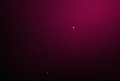 The primary subject of the image is a bright star in the night sky. The setting is a dark, pink background, which creates a contrasting and dramatic effect. The visual style is a photo, capturing the star and its surroundings. The colors and mood of the image are predominantly dark and moody, with the pink background adding a touch of warmth and contrast to the scene.