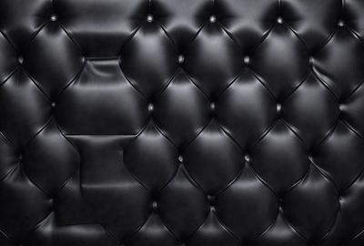 The primary subject of the image is a black leather couch. The setting is a black background, which creates a striking contrast with the couch. The visual style is a close-up of the couch, showcasing its texture and details. The colors and mood of the image are predominantly black, giving it a sleek and modern appearance.