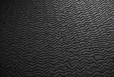 The primary subject of the image is a black, textured surface with a pattern resembling a car's tire. The setting is a dark room, and the visual style appears to be a close-up of the pattern. The colors and mood of the image are predominantly dark and moody, with the black surface and pattern creating a sense of depth and intrigue.