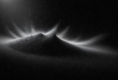 The primary subject of the image is a mountain of sand, which appears to be a black and white photo. The setting is a dark background, and the visual style is a close-up of the sand mountain. The colors and mood of the image are predominantly black and white, giving it a dramatic and artistic appearance.