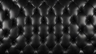 The primary subject of the image is a black leather couch. The setting is a black background, which creates a striking contrast with the couch. The visual style is a close-up of the couch, showcasing its texture and details. The colors and mood of the image are predominantly black, giving it a sleek and modern appearance.