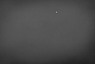 The primary subject of the image is a single white dot in the middle of a black background. The setting is a dark, empty sky, with no other objects or elements visible. The visual style is a simple, minimalist representation of the dot against the black background. The colors and mood are predominantly dark and moody, with the white dot being the only point of light in the scene.