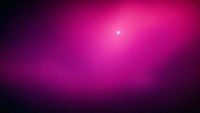 The primary subject of the image is a pink background with a bright pink sun in the center. The setting is a purple sky, which creates a contrasting backdrop for the sun. The visual style is a combination of a photo and digital art, as the sun appears to be a digital element placed in the middle of the purple sky. The colors and mood of the image are predominantly pink, creating a warm and inviting atmosphere.