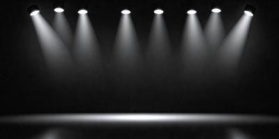 The primary subject of the image is a black background with a row of five lights shining on it. The lights are positioned at various heights and angles, creating a visually striking scene. The setting is a dark room with the lights as the main focus, and the visual style is a black and white photo. The overall atmosphere is dramatic and artistic, with the lights creating a sense of depth and dimension.
