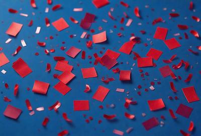 The primary subject of the image is a close-up of red confetti pieces scattered on a blue surface. The setting appears to be a room with a blue table or floor. The visual style is a close-up of the confetti, which gives a sense of depth and detail. The colors and mood of the image are predominantly red and blue, creating a festive and lively atmosphere.