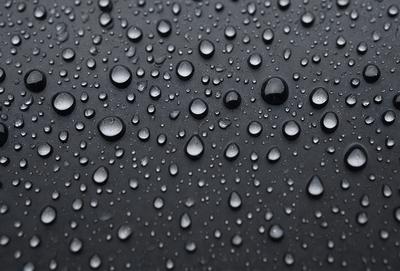 The primary subject of the image is a close-up of a rainy day, with a black background and a focus on the raindrops. The setting is a rainy day, and the visual style is a photo. The colors and mood of the image are dominated by the raindrops, which appear as tiny bubbles on the black surface. The overall atmosphere of the image is moist and atmospheric.
