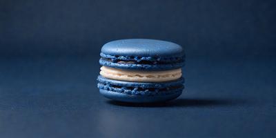 The primary subject of the image is a blue macaroon with white filling. The setting is a blue background, which creates a contrast with the blue and white colors of the macaroon. The visual style is a close-up of the macaroon, showcasing its details. The colors and mood of the image are predominantly blue and white, giving it a clean and fresh appearance.