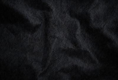 The primary subject of the image is a black fabric with a texture that resembles a woven or knitted material. The setting is a dark background, which emphasizes the fabric's texture and creates a moody atmosphere. The visual style is a close-up of the fabric, showcasing its intricate texture. The colors and mood of the image are predominantly dark, with the black fabric being the main focus.