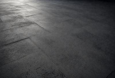 The primary subject of the image is a large, gray, concrete floor. The setting is a room with a tiled floor, and the visual style is a black and white photo. The colors and mood of the image are predominantly dark and moody, with the gray concrete floor and tiled walls creating a stark and minimalist atmosphere.