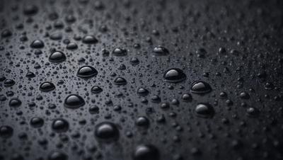 The primary subject of the image is a close-up of a black surface with numerous small droplets of water on it. The setting is a dark background, and the visual style appears to be a photo. The colors and mood of the image are predominantly dark, with the droplets of water being the main focus.