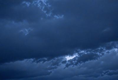 The primary subject of the image is a cloudy sky. The setting is a dark, cloudy day with a blue hue. The visual style is a photo of the sky, showcasing the clouds and their various shapes and sizes. The colors and mood of the image are predominantly dark and moody, with the blue hue adding a touch of calmness to the scene.
