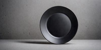 The primary subject of the image is a black plate. The plate is placed on a white surface, which serves as the background. The visual style of the image is a photo, capturing the plate's details and texture. The colors in the image are predominantly black and white, creating a simple and minimalist atmosphere.