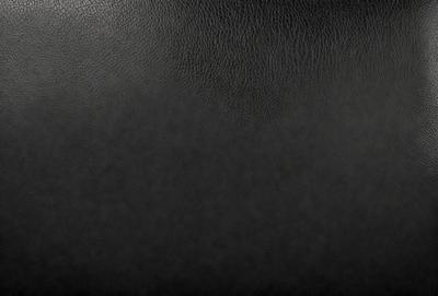 The primary subject of the image is a black leather seat. The setting is a black background, which creates a contrast with the leather seat. The visual style is a close-up of the leather seat, showcasing its texture and details. The colors and mood of the image are dark and moody, emphasizing the leather seat's richness and luxury.