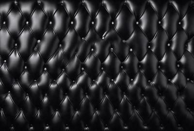 The primary subject of the image is a black leather couch. The setting is a black background, which creates a striking contrast with the couch. The visual style is a close-up of the couch, showcasing its texture and details. The colors and mood of the image are predominantly black, giving it a sleek and modern appearance.