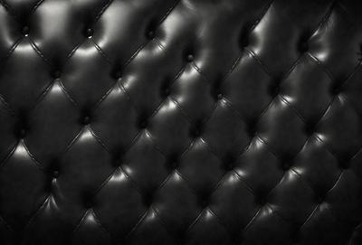 The primary subject of the image is a black leather couch. The setting is a black background, which creates a striking contrast with the couch. The visual style is a close-up of the couch, showcasing its texture and details. The colors and mood of the image are predominantly black, giving it a sleek and modern appearance.