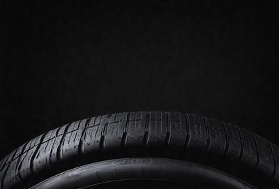 The primary subject of the image is a tire, which is the main object, scene, or focus of the image. The tire is black and is placed on a black background, giving it a dramatic and striking appearance. The visual style of the image is a close-up of the tire, emphasizing its details and texture. The colors and mood of the image are predominantly dark, with the black tire and background creating a moody and intense atmosphere.