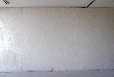 The image features a white wall with a large, empty space. The wall appears to be made of cement, giving it a textured and somewhat rough appearance. The primary subject of the image is the white wall, which serves as the main focus of the scene. The setting is a simple, empty room with no other objects or distractions visible. The visual style of the image is a close-up of the wall, emphasizing its texture and the emptiness of the space. The colors and mood of the image are pre