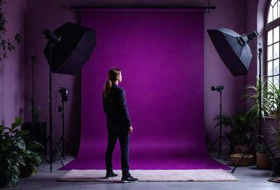 The primary subject of the image is a man standing in front of a purple background. He is wearing a black jacket and appears to be posing for a photo. The setting is a studio, with a pink background and a purple tarp. The visual style is a photo, and the colors and mood are predominantly purple and pink, creating a warm and inviting atmosphere.