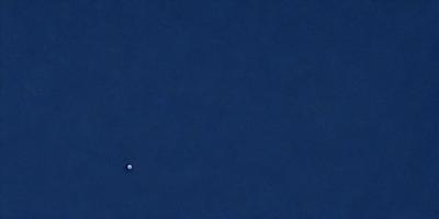 The primary subject of the image is a small white dot in the middle of a vast blue sky. The setting is a clear, cloudless day with no visible landmarks or objects. The visual style is a realistic representation of the scene, and the colors and mood are predominantly blue, creating a serene and peaceful atmosphere.