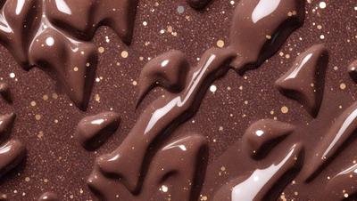 The primary subject of the image is a close-up of a chocolate-covered cake with a shiny, glittery surface. The cake is decorated with chocolate drizzles and gold sprinkles, giving it a luxurious and visually appealing appearance. The setting is a table, and the visual style is a close-up photo. The colors and mood of the image are predominantly brown and gold, creating a warm and inviting atmosphere.