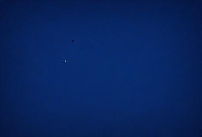 The primary subject of the image is the moon, which is seen in the sky. The setting is a dark blue sky, and the visual style is a photo. The colors and mood of the image are predominantly dark and moody, with the moon being the main focal point.