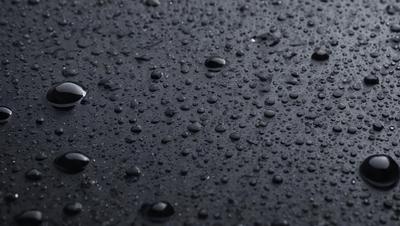 The primary subject of the image is a close-up of a black surface with numerous droplets of water on it. The setting is a dark background, and the visual style appears to be a photo. The colors and mood of the image are predominantly dark, with the droplets of water being the main focus.