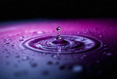 The primary subject of the image is a single drop of water, which is floating on a purple surface. The setting appears to be a dark, possibly pink, background. The visual style of the image is a close-up of the drop of water, emphasizing its details. The colors and mood of the image are predominantly purple, creating a calm and serene atmosphere.