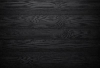 The primary subject of the image is a wooden wall with a black background. The setting is a room with a wooden floor. The visual style is a close-up of the wooden wall, showcasing its texture and details. The colors and mood of the image are dark and moody, with the black wooden wall and floor creating a dramatic atmosphere.