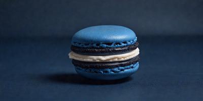The primary subject of the image is a blue and white macaron, which is sitting on a blue surface. The setting appears to be a table or countertop. The visual style of the image is a close-up of the macaron, showcasing its details. The dominant colors in the image are blue and white, creating a clean and fresh atmosphere.