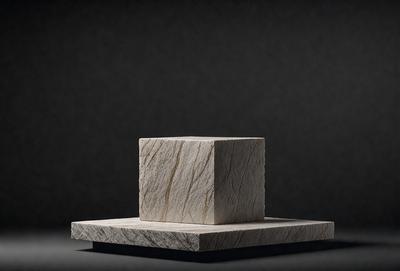 The primary subject of the image is a square block of marble, which is the main object in the scene. The setting is a dark background, which contrasts with the marble block. The visual style is a close-up of the marble block, showcasing its texture and details. The colors and mood of the image are predominantly dark, with the marble block being the main focal point.