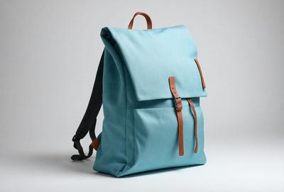 The primary subject of the image is a blue backpack with a brown strap. The backpack is placed on a white background, which creates a clean and minimalist appearance. The visual style of the image is a photo, capturing the details of the backpack and its strap. The colors and mood of the image are predominantly blue and white, giving it a calm and serene atmosphere.