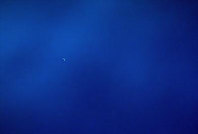 The primary subject of the image is the moon, which is visible in the sky. The setting is a dark blue sky, which creates a contrasting backdrop for the moon. The visual style of the image is a photo, capturing the moon in its natural environment. The colors and mood of the image are predominantly dark and moody, with the blue sky and the moon creating a sense of depth and tranquility.