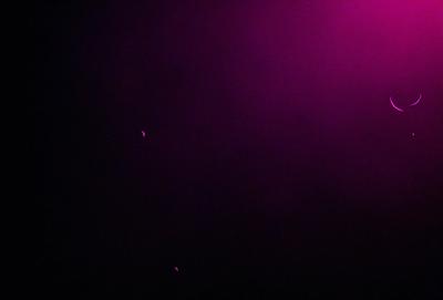 The image is a black and white photo of a purple background. The primary subject is a bright light source, which appears to be a light bulb. The light source is located in the upper left corner of the image. The setting is a dark room, and the visual style is a classic black and white photo. The colors and mood of the image are predominantly purple, creating a moody and dramatic atmosphere.
