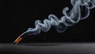 The primary subject of the image is a lit cigarette, which is the main focus of the scene. The setting is a dark room, with the cigarette smoke curling upwards, creating a dramatic and moody atmosphere. The visual style of the image is a close-up of the lit cigarette, emphasizing its glow and the smoke it emits. The colors in the image are predominantly dark, with the lit cigarette being the only source of light, creating a sense of intensity and drama