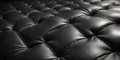 The primary subject of the image is a black leather couch. The setting is a room with a black background, which creates a contrast with the couch. The visual style is a close-up of the couch, showcasing its texture and details. The colors and mood of the image are predominantly black, giving it a sleek and modern appearance.