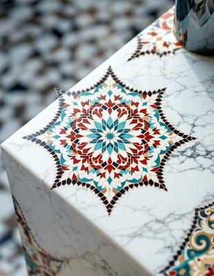 The primary subject of the image is a decorative tile with a colorful design, featuring a flower pattern. The tile is placed on a marble table, which serves as the setting for the scene. The visual style of the image is a close-up of the tile, showcasing its intricate details. The colors and mood of the image are vibrant and lively, with the flower pattern adding a touch of natural beauty to the overall atmosphere.