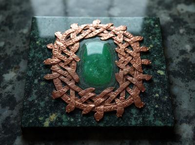 The primary subject of the image is a green stone with a metal design, possibly a copper or brass piece, surrounding it. The setting is a table, and the visual style appears to be a combination of a photo and a digital art or illustration. The colors in the image are predominantly green and brown, giving the scene a natural and earthy atmosphere.