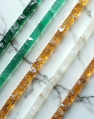 The primary subject of the image is a row of five different colored glass sticks, each with a unique hue, arranged on a white marble countertop. The setting is a clean and elegant environment, with the marble countertop serving as the background. The visual style of the image is a close-up of the glass sticks, showcasing their vibrant colors and unique designs. The colors and mood of the image are predominantly white and clean, with the vibrant colors of the glass sticks adding