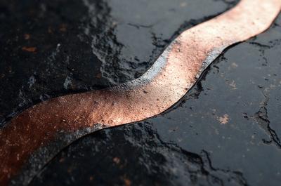 The primary subject of the image is a piece of copper wire, which is lying on a black surface. The setting appears to be a concrete or asphalt surface, possibly a road or a sidewalk. The visual style of the image is a close-up of the copper wire, showcasing its texture and color. The dominant colors in the image are black and copper, creating a contrast between the two. The overall atmosphere of the image is somewhat mysterious and intriguing, as the viewer is drawn to the