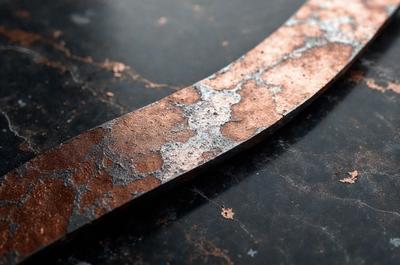 The primary subject of the image is a piece of metal, possibly a metal plate or a metal bar, with a rusty appearance. The metal is placed on a black surface, which could be a table or a countertop. The visual style of the image is a close-up of the metal, showcasing its texture and rusty appearance. The colors in the image are predominantly black and brown, giving the scene a somewhat moody and aged appearance.