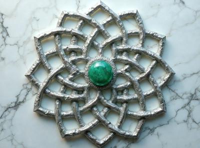 The primary subject of the image is a silver metal art piece featuring a green gemstone in the center. The setting is a marble surface, and the visual style appears to be a combination of digital art and illustration. The colors in the image are predominantly silver and green, creating a sense of elegance and sophistication.