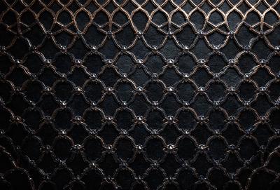 The primary subject of the image is a patterned surface, which appears to be a black background with a metallic texture. The setting is a dark room, and the visual style is a close-up of the patterned surface. The colors and mood of the image are predominantly dark and moody, with the metallic texture adding a sense of depth and intricacy to the pattern.